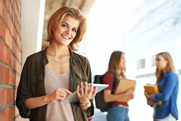 Technologia pomaga mi się wyróżniać na studiach. Portret uśmiechniętej studentki uniwersytetu stojącej na kampusie za pomocą tabletu cyfrowego z przyjaciółmi w tle. - Zdjęcie, obraz