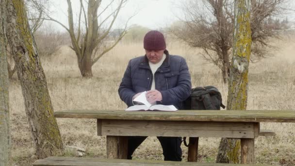 człowiek w naturze, odwraca stronę i czyta grubą książkę siedzącą przy drewnianym stole - Materiał filmowy, wideo