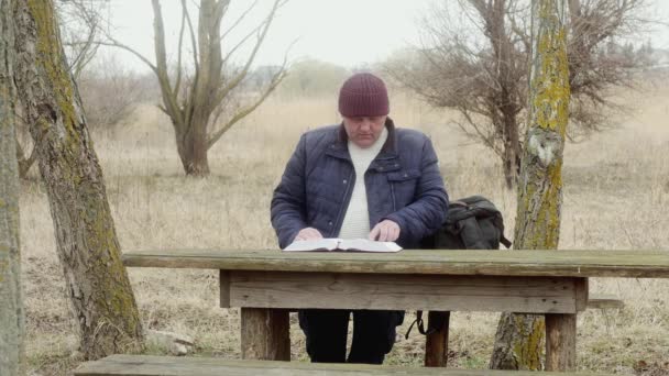  mężczyzna na zewnątrz czyta książkę z zainteresowaniem siedzącą przy drewnianym stole między drzewami - Materiał filmowy, wideo
