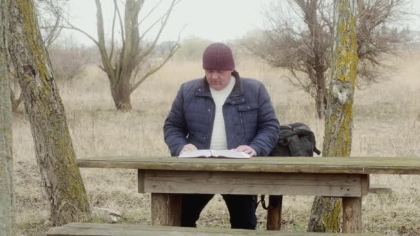 człowiek na zewnątrz czyta grubą książkę siedzącą przy drewnianym stole między drzewami. - Materiał filmowy, wideo