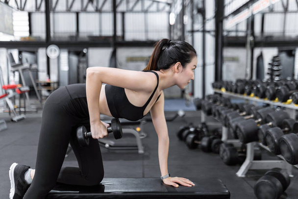 exercise concept The female exercise beginner doing dumbbell workout by resting her left knee and left hand on the bench and raise her right elbow up with a dumbbell in her hand. - Photo, image