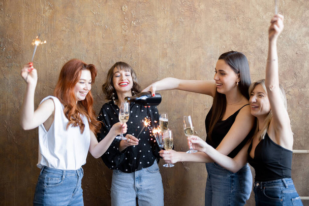 Přítelkyně pijící šumivé víno a pálící šumáky, oslavující společně uvnitř - Fotografie, Obrázek