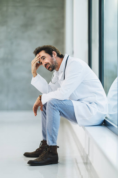 Κουρασμένος γιατρός κάνει ένα γρήγορο διάλειμμα και σκεπτικός κάθεται σε ένα άδειο διάδρομο του νοσοκομείου. - Φωτογραφία, εικόνα