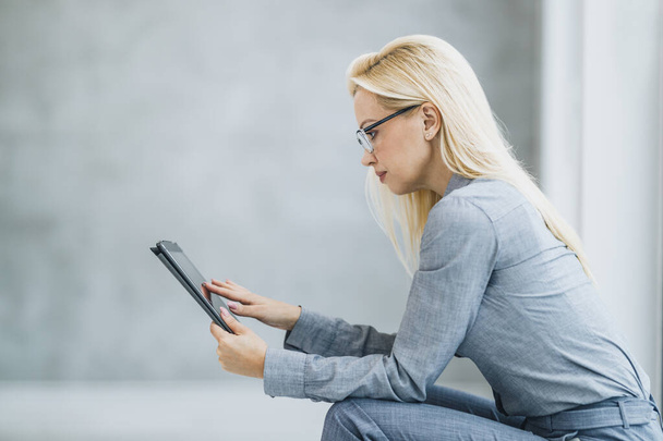 Pewna siebie bizneswoman korzystająca z tabletu cyfrowego siedząc przy oknie w biurze podczas przerwy. - Zdjęcie, obraz