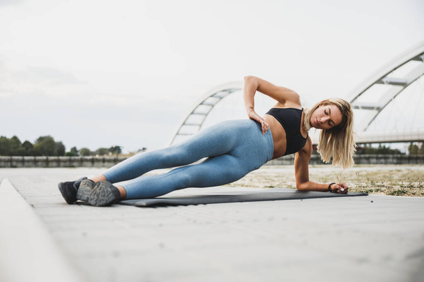 Young fit woman doing side plank exercises  during working up outdoors, on riverside city promenade. - Fotó, kép
