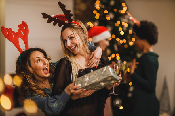 Wesołe przyjaciółki przytulające się i dające sobie prezenty podczas spotkania w domu, aby świętować Boże Narodzenie lub Nowy Rok. - Zdjęcie, obraz