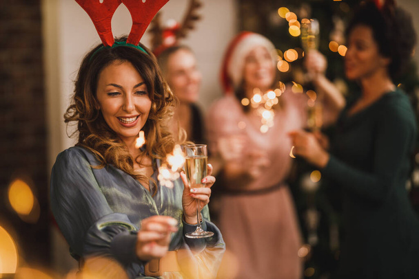 Mujer joven sonriente brindando con champán y jugando con bengalas mientras celebra la Navidad o Año Nuevo con sus amigos en la fiesta en casa. - Foto, imagen