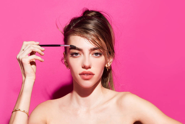 Combs eyebrows with a brush in a beauty salon. Woman with long eyelashes and thick eyebrows. Woman portrait isolated on pink studio background. Natural beauty brows. - Φωτογραφία, εικόνα