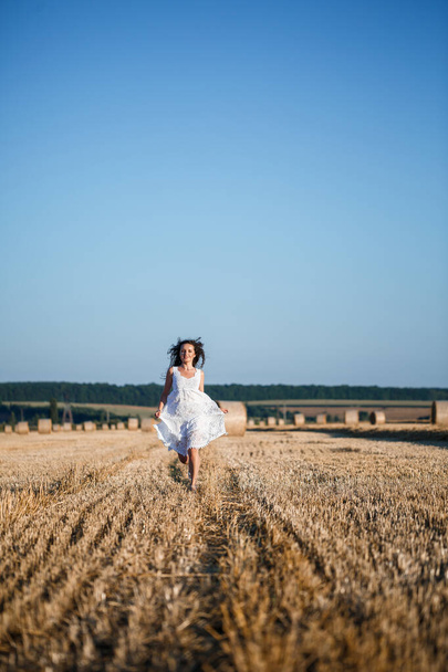 A young beautiful woman in a white summer dress stands on a mown wheat field with huge sheaves of hay, enjoying nature. Nature in the village. Selective focus - Fotoğraf, Görsel