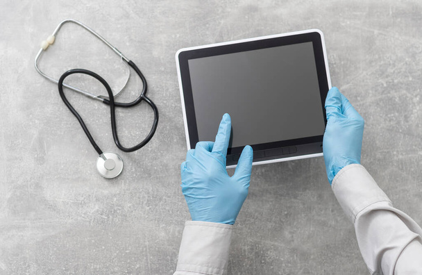 Κάτοψη των χεριών του γιατρού με τον υπολογιστή tablet. Καρδιακή εξέταση στο νοσοκομείο. Θεραπευτής κάθεται στο γραφείο με στηθοσκόπιο, ιατρικά γάντια. Εξέταση και συμβουλευτική στην κλινική - Φωτογραφία, εικόνα