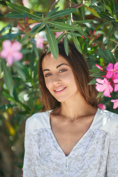 Pretty woman with perfect skin and charming smile near blooming pink flowers in garden - Foto, afbeelding