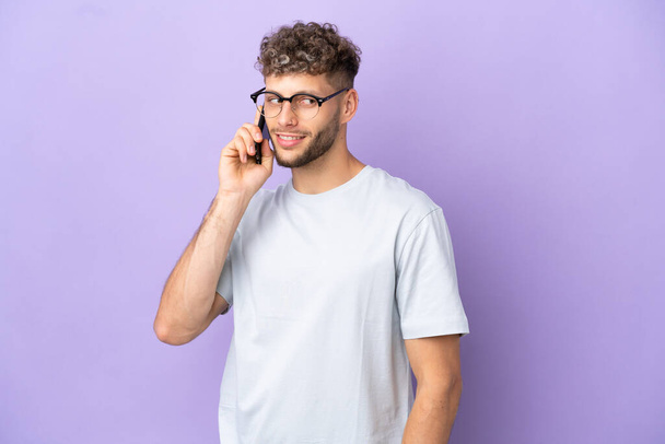 Delivery caucasian man isolated on purple background keeping a conversation with the mobile phone with someone - Photo, image
