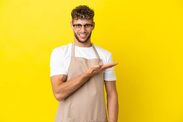 Restaurant Kellner blonder Mann isoliert auf gelbem Hintergrund präsentiert eine Idee, während er lächelnd in Richtung - Foto, Bild