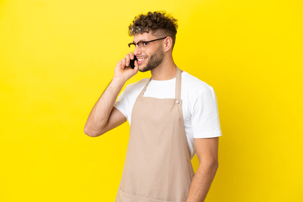 Restauracja kelner blondynka człowiek odizolowany na żółtym tle prowadzenie rozmowy z telefonu komórkowego z kimś - Zdjęcie, obraz