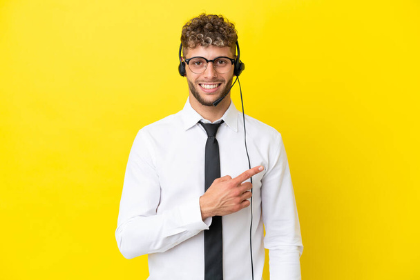 Telemarketer blonde Mann arbeitet mit einem Headset isoliert auf gelbem Hintergrund zeigt auf die Seite, um ein Produkt zu präsentieren - Foto, Bild