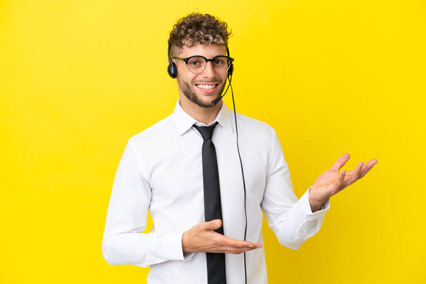 Telemarketer blonde man working with a headset isolated on yellow background extending hands to the side for inviting to come - Zdjęcie, obraz