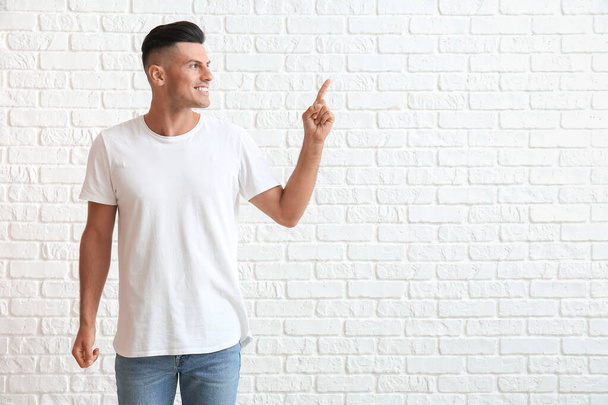 Όμορφος νεαρός άνδρας με κομψό t-shirt δείχνει κάτι σε λευκό φόντο τούβλο - Φωτογραφία, εικόνα