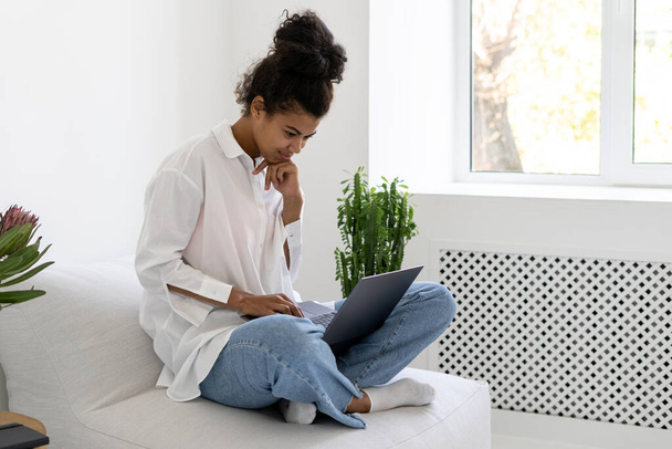 Vista lateral da bela senhora afro-americana com laptop surf web na acolhedora sala de estar em estilo nórdico. Comunicação online por videochamada durante a pandemia de covid-19 - Foto, Imagem