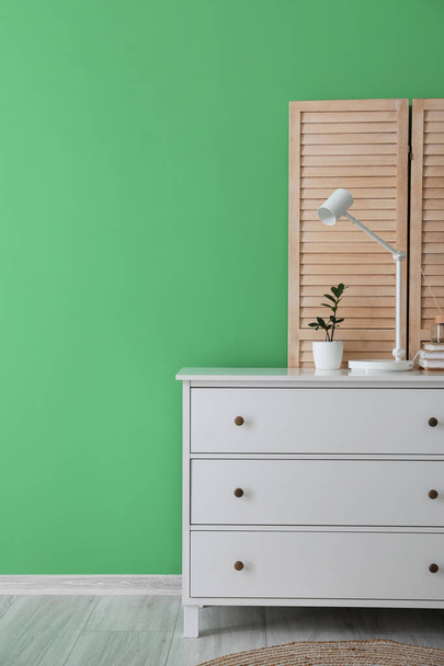 Cómoda con lámpara blanca, maceta y pantalla plegable cerca de la pared verde - Foto, Imagen