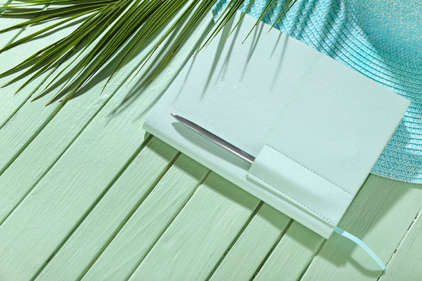 Стильный ноутбук с ручкой, шляпой и пальмовым листком на зеленом деревянном фоне - Фото, изображение