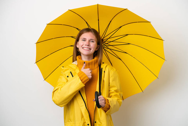 Junge Engländerin mit regenfestem Mantel und Regenschirm auf weißem Hintergrund, die eine Daumen-hoch-Geste macht - Foto, Bild