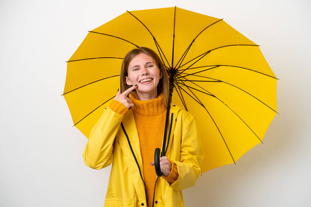 Jonge Engelse vrouw met regenbestendige jas en paraplu geïsoleerd op witte achtergrond geven een duim omhoog gebaar - Foto, afbeelding