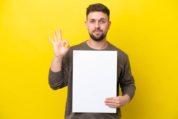 Junger kaukasischer Mann isoliert auf gelbem Hintergrund mit leerem Plakat und OK-Zeichen - Foto, Bild