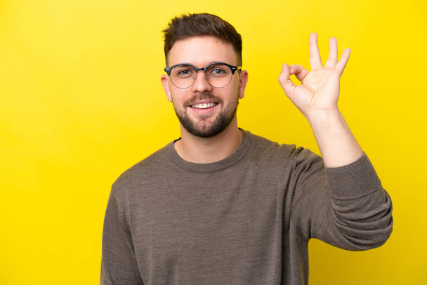Young caucasian man isolated on yellow background With glasses and doing OK sign - Photo, Image