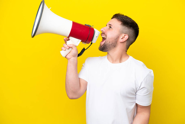 Young caucasian man isolated on yellow background shouting through a megaphone to announce something in lateral position - Fotografie, Obrázek