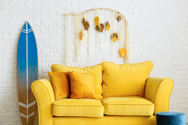 Cómodo sofá, puf y tabla de surf cerca de la pared de ladrillo blanco - Foto, imagen