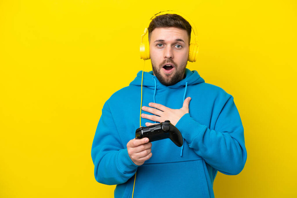 Giovane uomo caucasico che gioca con un controller di videogiochi isolato su sfondo giallo sorpreso e scioccato mentre guarda a destra - Foto, immagini