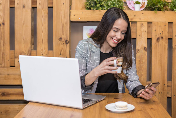 Positive junge Remote-Arbeiterin trinkt Getränke, während sie während der Online-Arbeit am Netbook im Café soziale Medien auf dem Handy scrollt - Foto, Bild