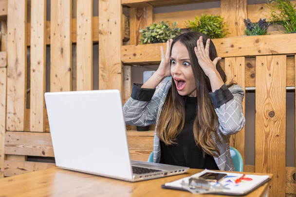 Geschockte Millennial-Arbeiterin hält die Hände auf dem Kopf, während sie empfangene E-Mails mit unglaublichen Neuigkeiten auf Netbook am bequemen Arbeitsplatz liest - Foto, Bild