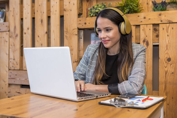Jeune femme écoutant de la musique dans les écouteurs sans fil et tapant sur netbook à table avec smartphone et presse-papiers pendant la journée de travail à distance - Photo, image