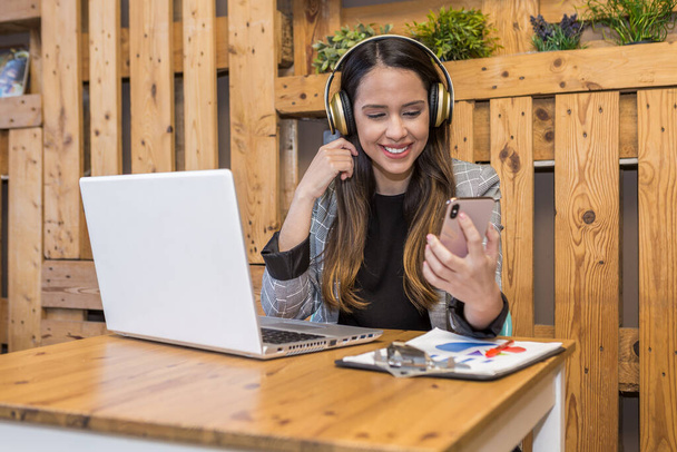 Positive Remote-Arbeiterin in drahtlosen Kopfhörern, die am Holztisch sitzt und während der Online-Arbeit am Netbook Nachrichten auf dem Handy checkt - Foto, Bild