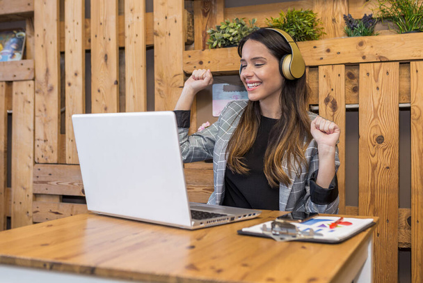 Onnellinen nuori nainen kauko asiantuntija muodollinen kulumista kuunnella musiikkia kuulokkeet ja puristamalla nyrkit samalla riemuiten saavutettu tuloksia työn netbook pöydässä älypuhelimella ja leikepöydällä - Valokuva, kuva