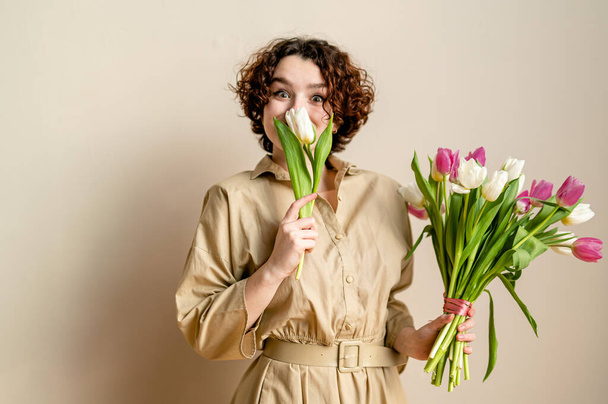 portrait d'une fille bouclée souriante avec des tulipes sur un fond beige dans une robe beige. espace de copie, palette de couleurs pastel - Photo, image
