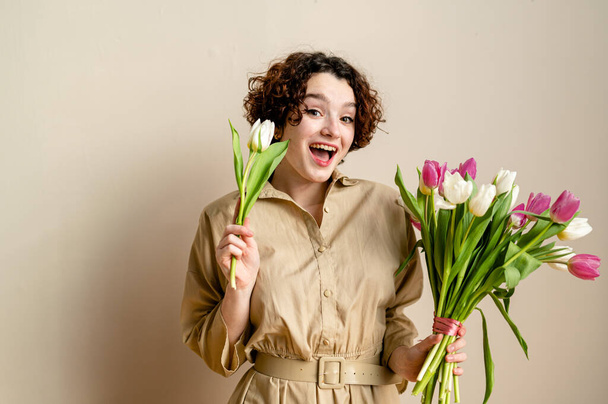 portrait of a smiling curly girl with tulips on a beige background in a beige dress. copy space, pastel color palette - Photo, Image
