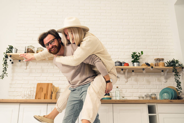 caucasian middle-aged bearded man having fun and lifting his fiancee piggyback in their modern bright kitchen - Photo, Image
