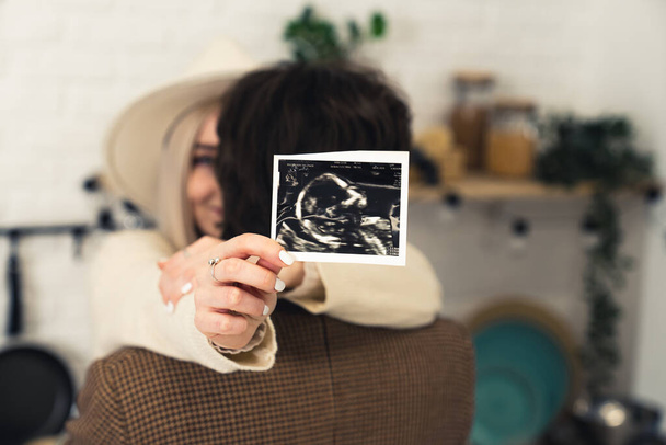Glückliches Paar umarmt, kaukasische Frau mit Ultraschallbild ihres Babys, Fokus auf Vordergrund - Foto, Bild