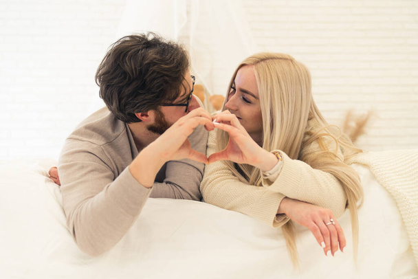 Cute Caucasian Couple Looking At Each Other Forming Heart Shape With Their Palms While Laying On Their Bed - Photo, Image