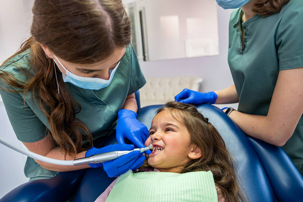 Özel kıyafetli genç dişçiler dişçi koltuğunda oturan küçük bir kızın dişlerini tedavi ediyorlar. Dişçilik ve çocuk kavramı - Fotoğraf, Görsel
