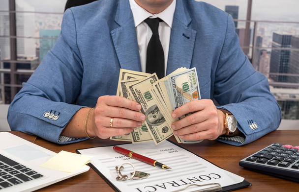 zakenman in een strak pak met dollars in zijn handen tekent een belangrijk zakelijk contract. Bedrijfs- en contractconcept - Foto, afbeelding