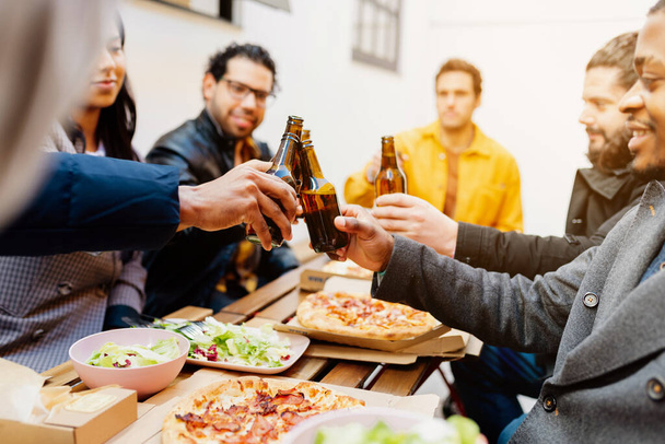πολυφυλετική ομάδα φίλων που κάνουν πρόποση με μπύρα κατά τη διάρκεια του εορτασμού ενός γεύματος σε βεράντα. Γυναίκες και άνδρες συνάδελφοι που τρώνε μετά την εργάσιμη ημέρα - Φωτογραφία, εικόνα