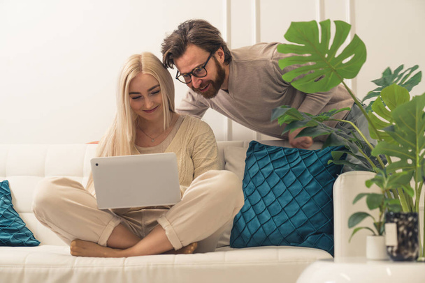 ελκυστικούς χαρούμενους παντρεμένους συζύγους που κάθονται σε έναν καναπέ, αγοράζουν δώρα και ψωνίζουν online χάρη στο ολοκαίνουργιο φορητό υπολογιστή τους - Φωτογραφία, εικόνα
