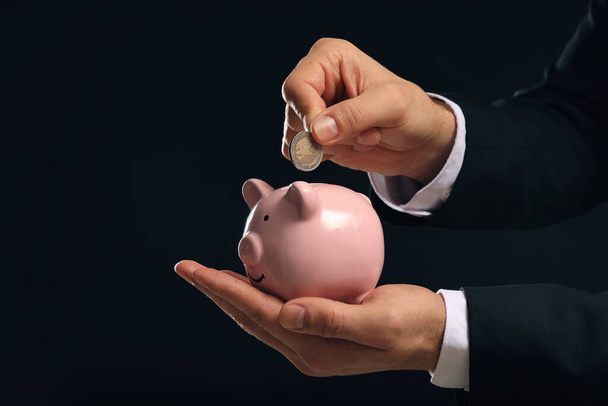 Businessman putting coin in piggy bank on black background, closeup - Foto, imagen