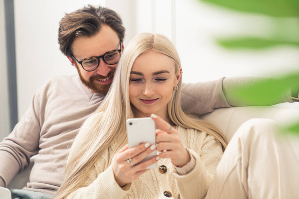 Ευρωπαϊκό ζευγάρι χιλιετιών που αναζητούν χαρούμενα σε ένα smartphone, ενώ κάθονται μαζί σε έναν καναπέ - Φωτογραφία, εικόνα