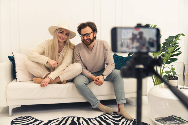 mladý kavkazský vlivný pár vlogování s chytrým telefonem, zatímco sedí na bílé pohovce v jejich moderním bytě - Fotografie, Obrázek