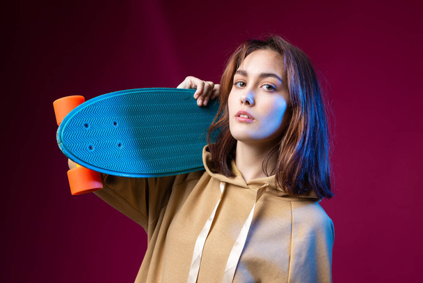 Young beautiful fashionable hipster girl dressed in a hoodie holds a skateboard in her hands on a purple studio background. - Foto, Imagem