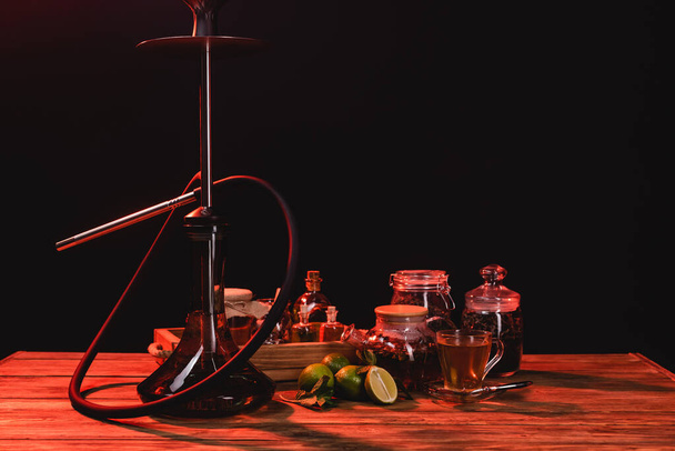 Chá, limas frescas e narguilé na mesa de madeira isolada em preto com luz vermelha - Foto, Imagem